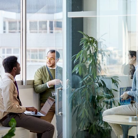 Coworkers talking in a room in an office. One person is seated at a table with an open laptop while the other is listening, behind the glass door there is also another employee visible in the conversation.