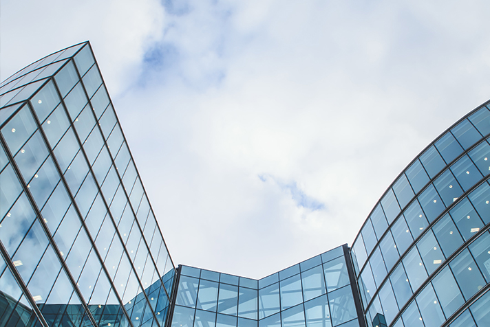 The top of a large modern glass building reflecting the clouded sky. The lights from inside the buildings are visible, suggesting offices within the buildings.