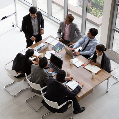 A birds eye view image of seven professionals gathered around a rectangular meeting table. They all are dressed in business attire, with most wearing suits or formal wear. Laptops, documents and coffee cups are visible on the table, suggesting an active work environment.