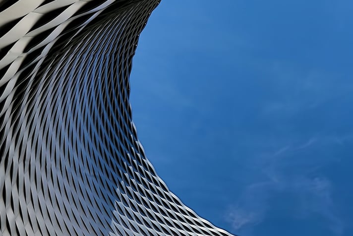 A curved modern building. The exterior has a weaving pattern made of a silver material. The sky in the background is bright and clear causing shadows on the exterior of the building.